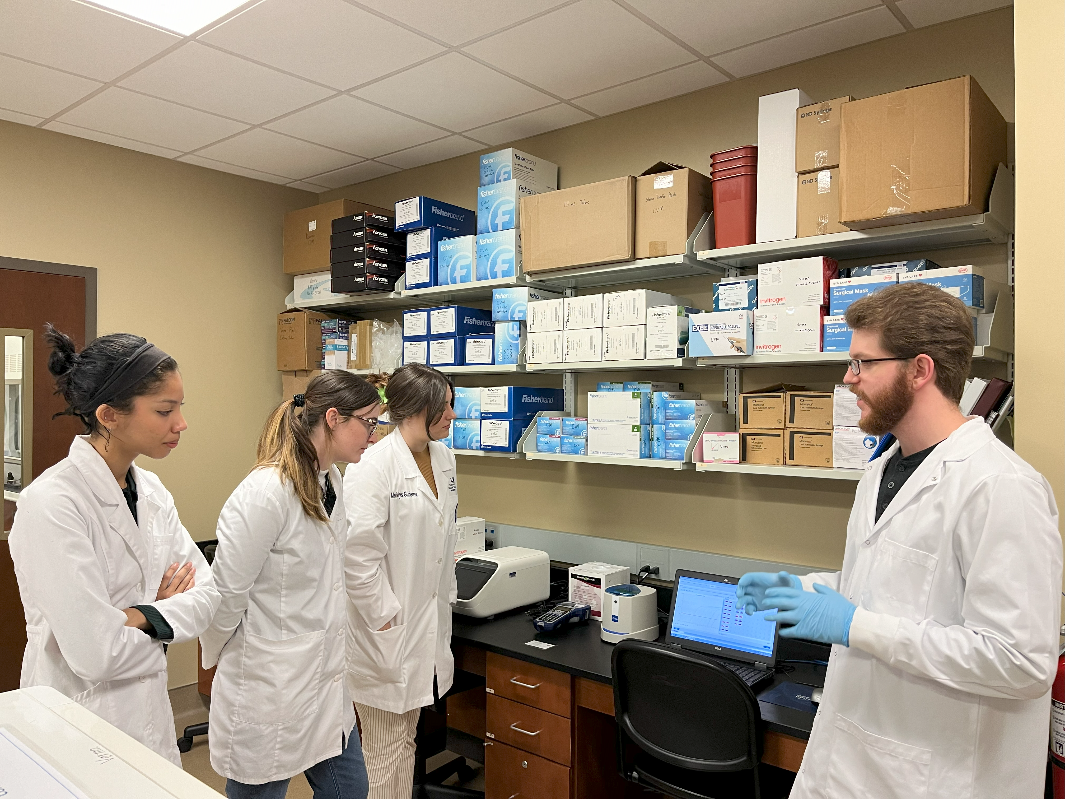 A group of 3 students and an instructor wearing lab coats in a lab